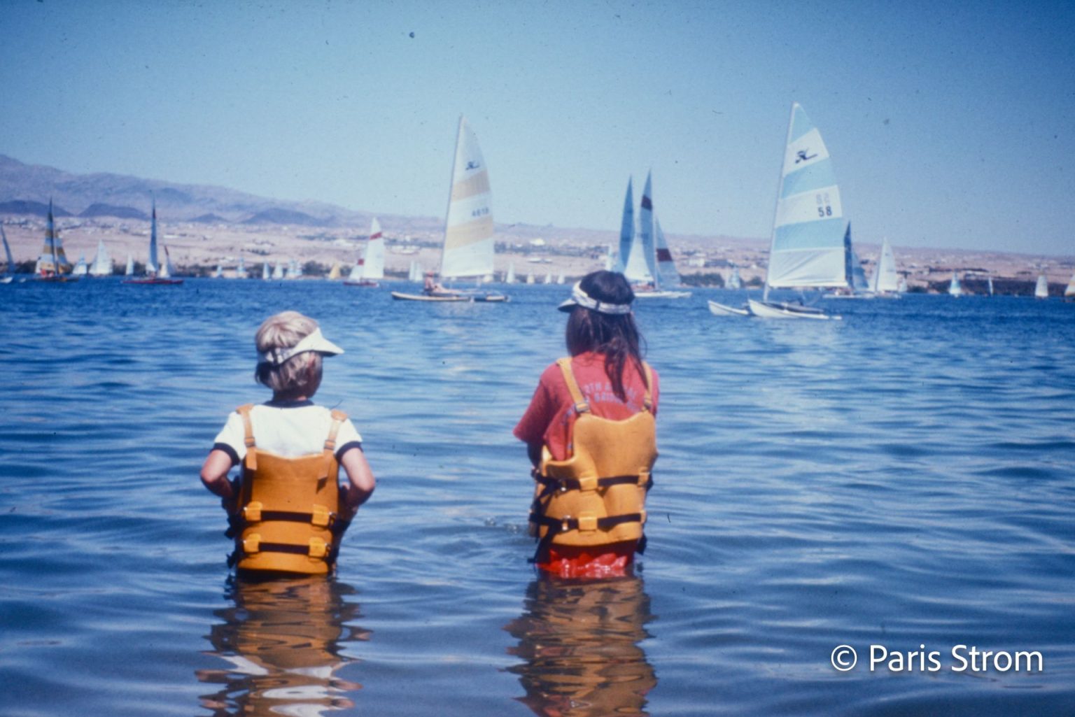 Two young children stand waist-deep in the water wearing lifejackets and watching the sailboats.