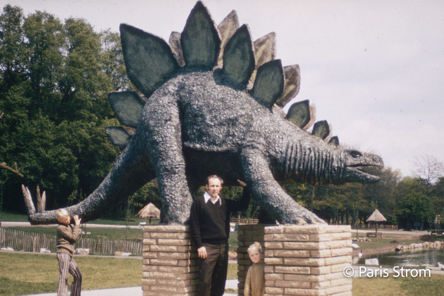 A man poses with a dinosaur statue and a young boy stands by the dinosaur's tail.