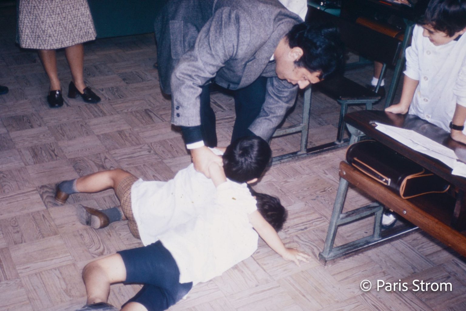Two toddler boys are scuffling on the ground and being separated by an adult male