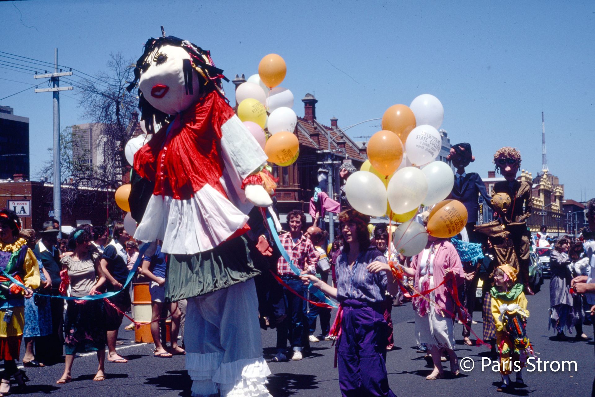 A parade showing a large puppet doll and people wearing costumes and holding balloons.