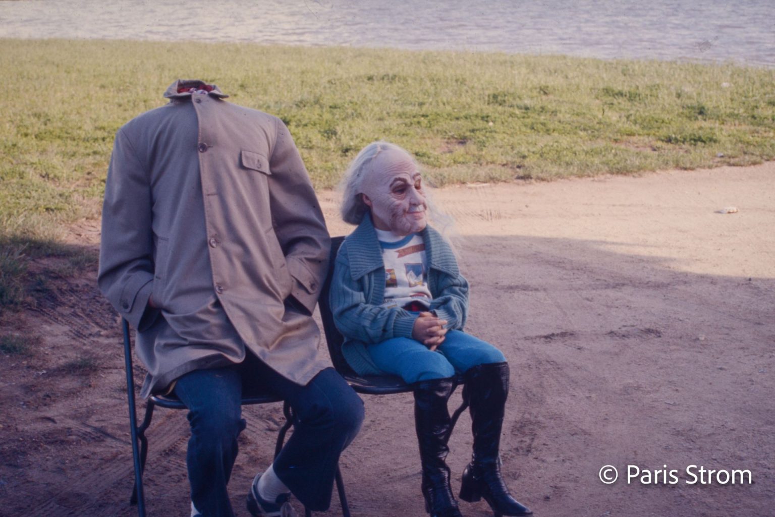 Two people sit on a bench. One has a coat buttoned over his head so he appears headless, and the child sitting next to him is wearing an old man mask.