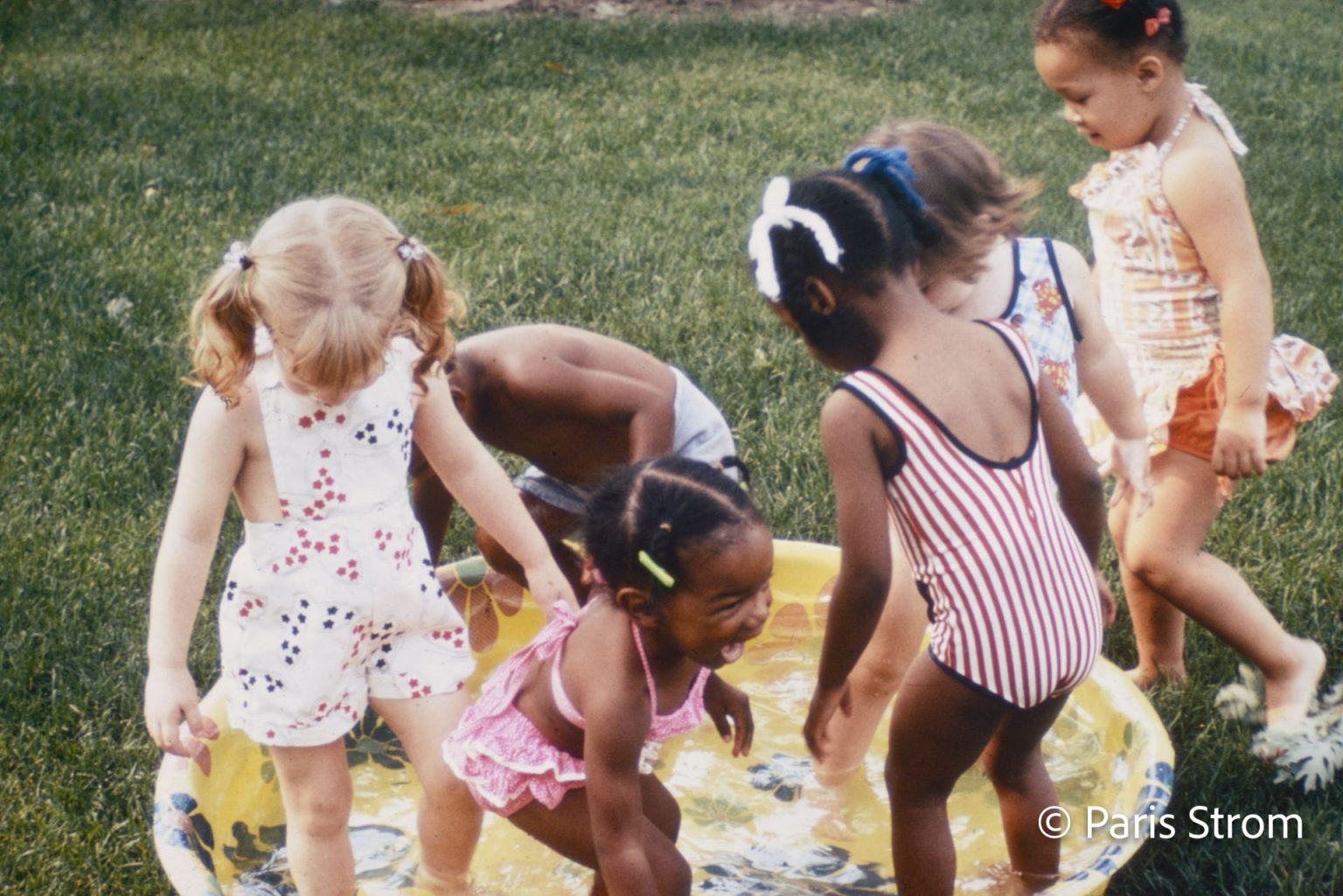 Young girls in swimsuits play in a kiddie pool on the grass.